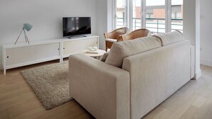 A modern living room with a beige sofa, a white entertainment unit, a flat-screen TV, wicker chairs, and a large window with a view of buildings.
