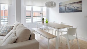 A modern living room with a beige sofa, white dining table, and chairs. There is a painting on the wall and a window with a view of buildings.
