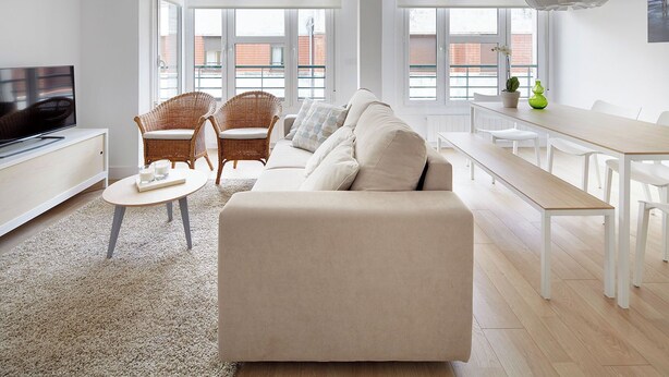 A modern living room with a sofa, two wicker chairs, a coffee table, a TV, and a dining area with a bench and chairs.