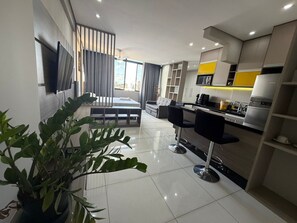 A modern kitchen with a dining area, a TV mounted on the wall, and a large window with curtains.