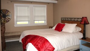 Een slaapkamer met een bed, een deken met een rood-wit patroon, een klok, een raam met jaloezieën, een nachtkastje met een lamp en een houten hoofdbord.