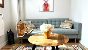 Ein Wohnzimmer mit grauem Sofa, gemusterten Kissen, einem runden Holztisch mit einer Vase weißer Blumen, einer schwarzen Stehlampe und einem Wandbild von einem Hirsch.