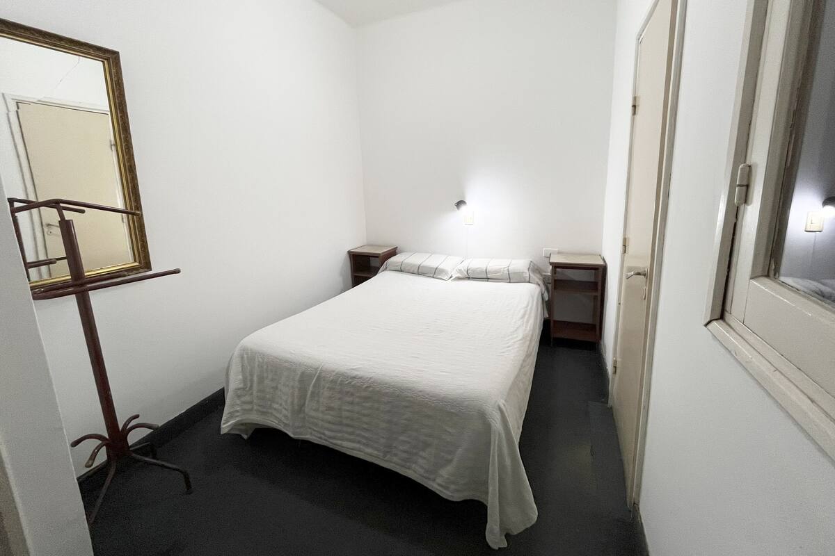 Ein kleines, schmales Hotelzimmer mit einem Einzelbett, Nachttischen, einem Spiegel und einem Fenster.