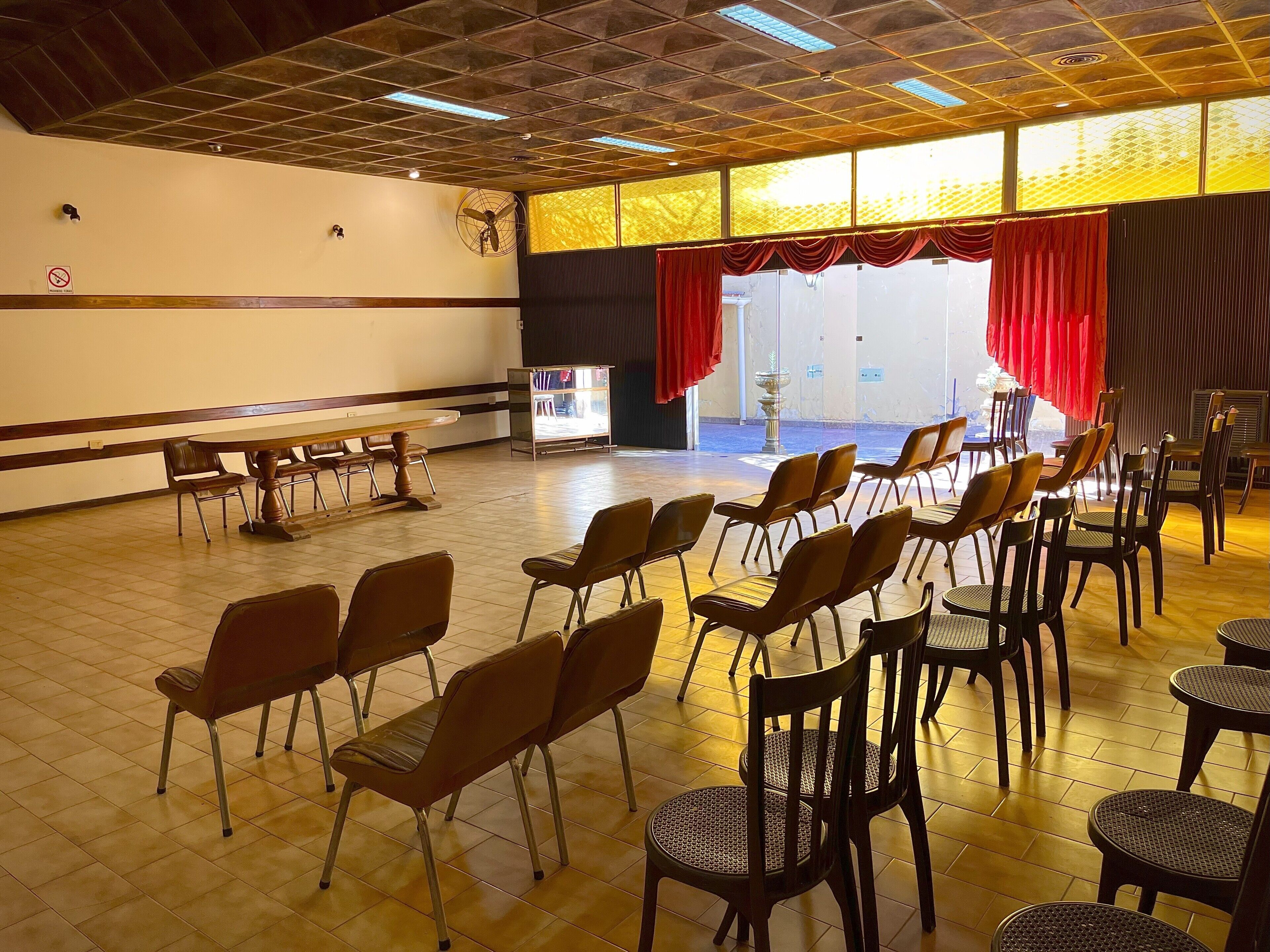 A spacious hall with rows of chairs and tables, a stage with red curtains, and a large window allowing natural light.