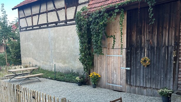 A rustic building with a red-tiled roof, wooden walls, and a wooden door. There is a wooden fence in front, and a table and chair set outside. - 60 sqm apartment on the ground floor with garden and courtyard for 2 people (Michelau in Oberfranken)