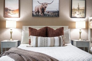 A bed with a white quilt and two brown decorative pillows, flanked by two bedside tables with lamps, in a room with a textured wall and framed pictures above the bed. - Hot Tub + Outdoor Kitchen: Stylish Home in Bryan (Bryan)