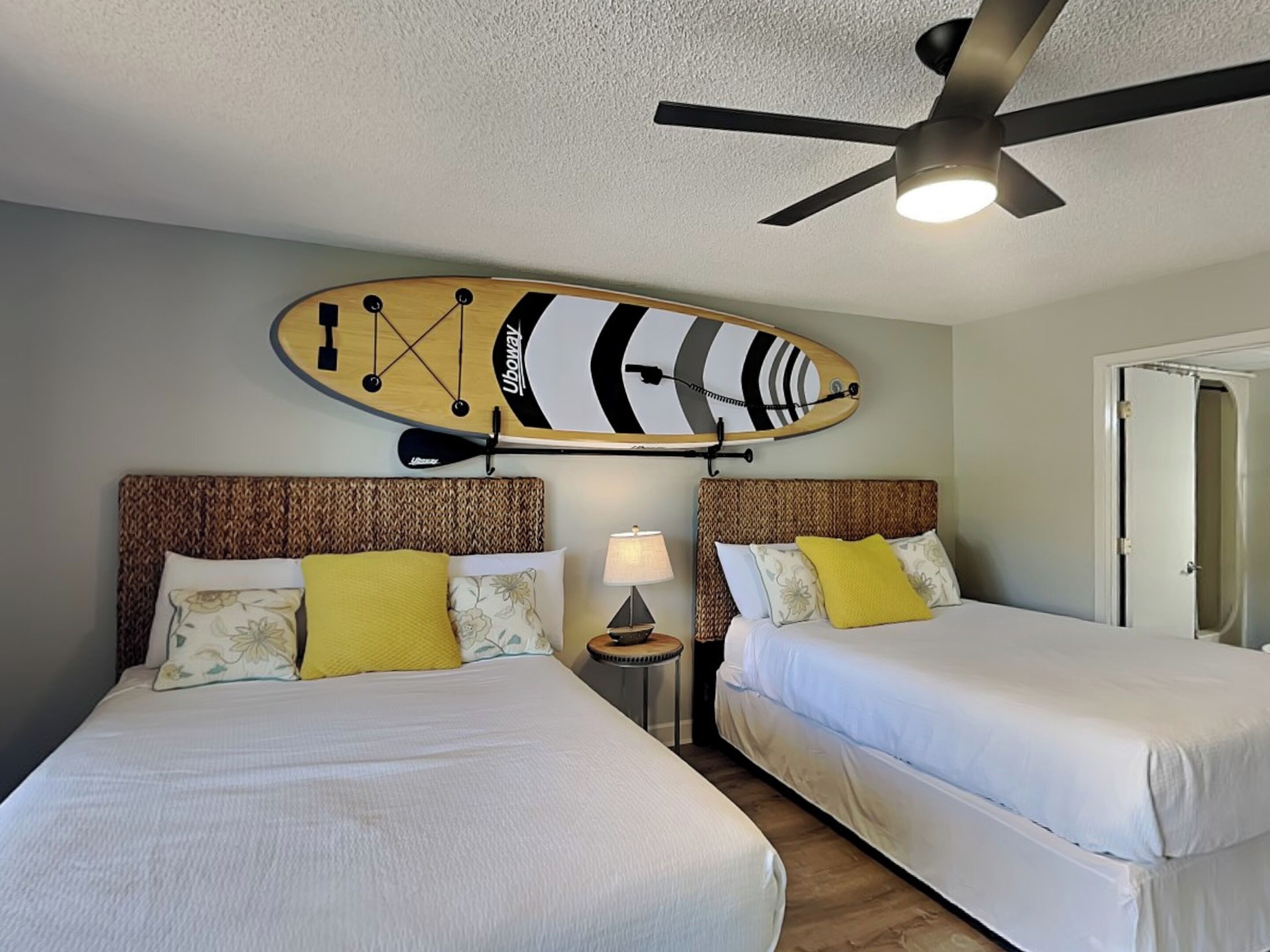 A hotel room with two beds, a ceiling fan, a wall-mounted surfboard, and a nightstand with a lamp.