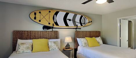 A hotel room with two beds, a ceiling fan, a wall-mounted surfboard, and a nightstand with a lamp.