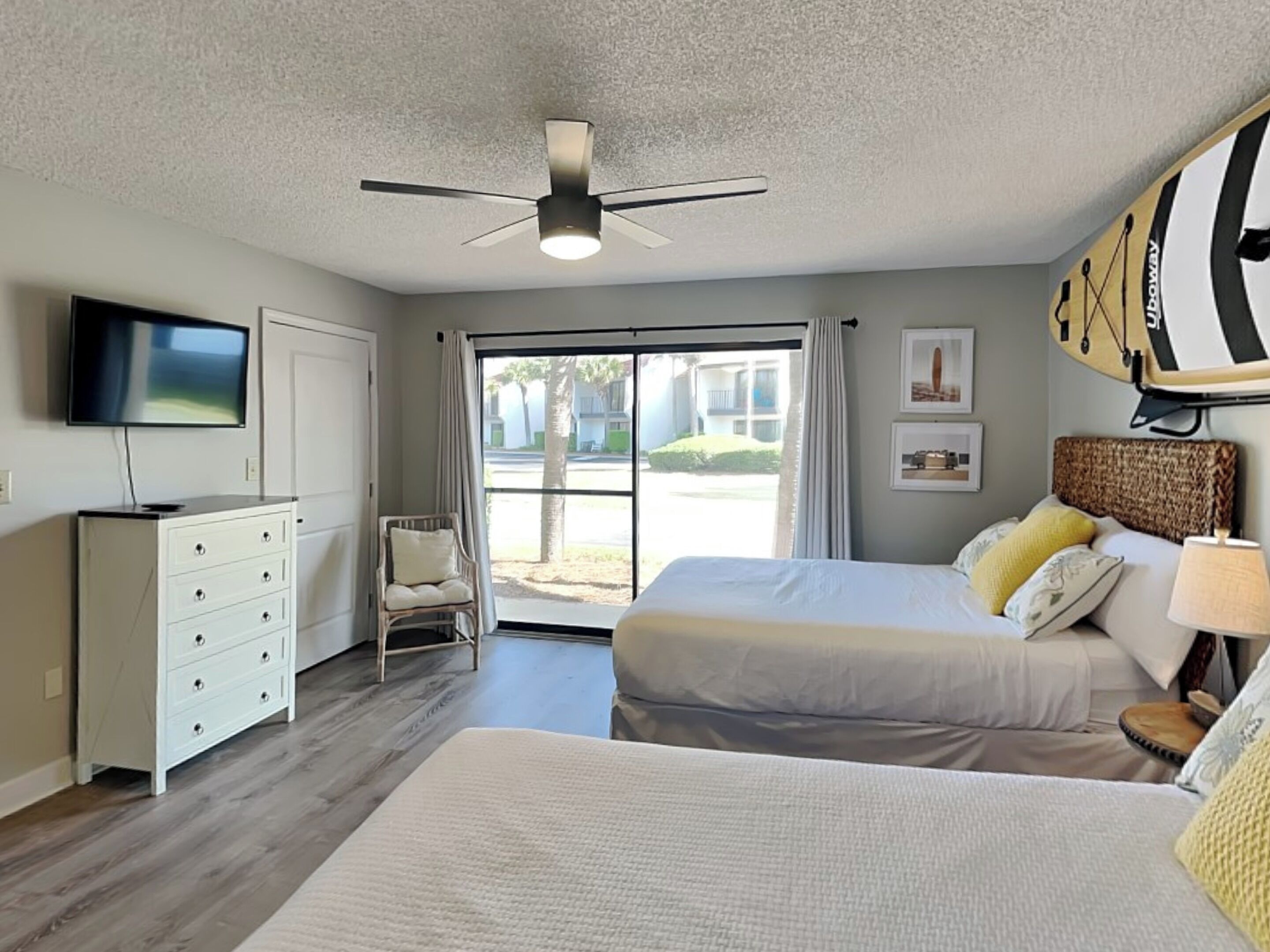A hotel room with a bed, a chair, a TV, a ceiling fan, and a view of the outside.