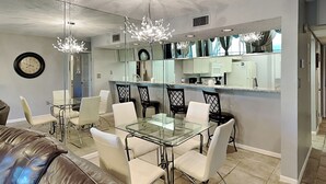 A modern dining area with a glass table and chairs, a chandelier, a clock, and a kitchen visible through a glass partition. - Edgewater Villa #2005 by Resort Collection (Panama City Beach)