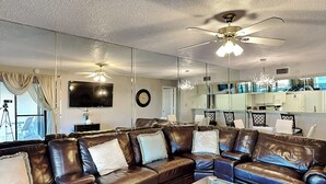 A modern living room with leather sofas, a glass coffee table, a ceiling fan, a flat-screen TV, and a kitchen area visible through a glass partition. - Edgewater Villa #2005 by Resort Collection (Panama City Beach)
