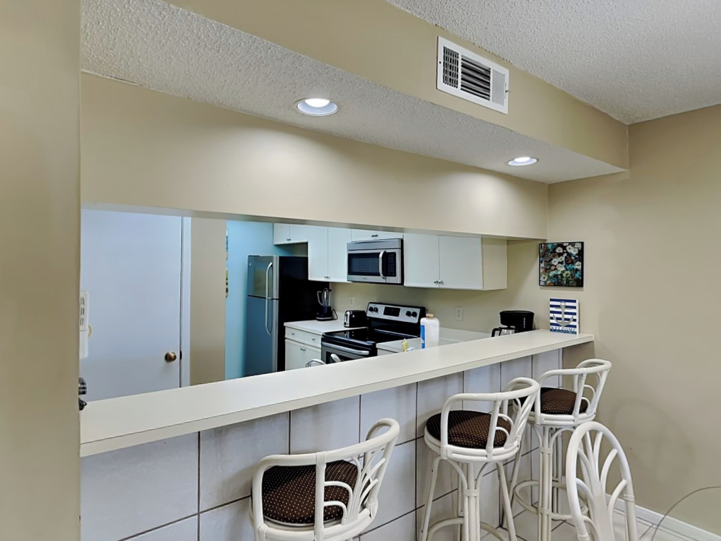 Una cocina con armarios blancos, microondas, refrigerador y estufa. Hay tres taburetes blancos con asientos de mimbre.