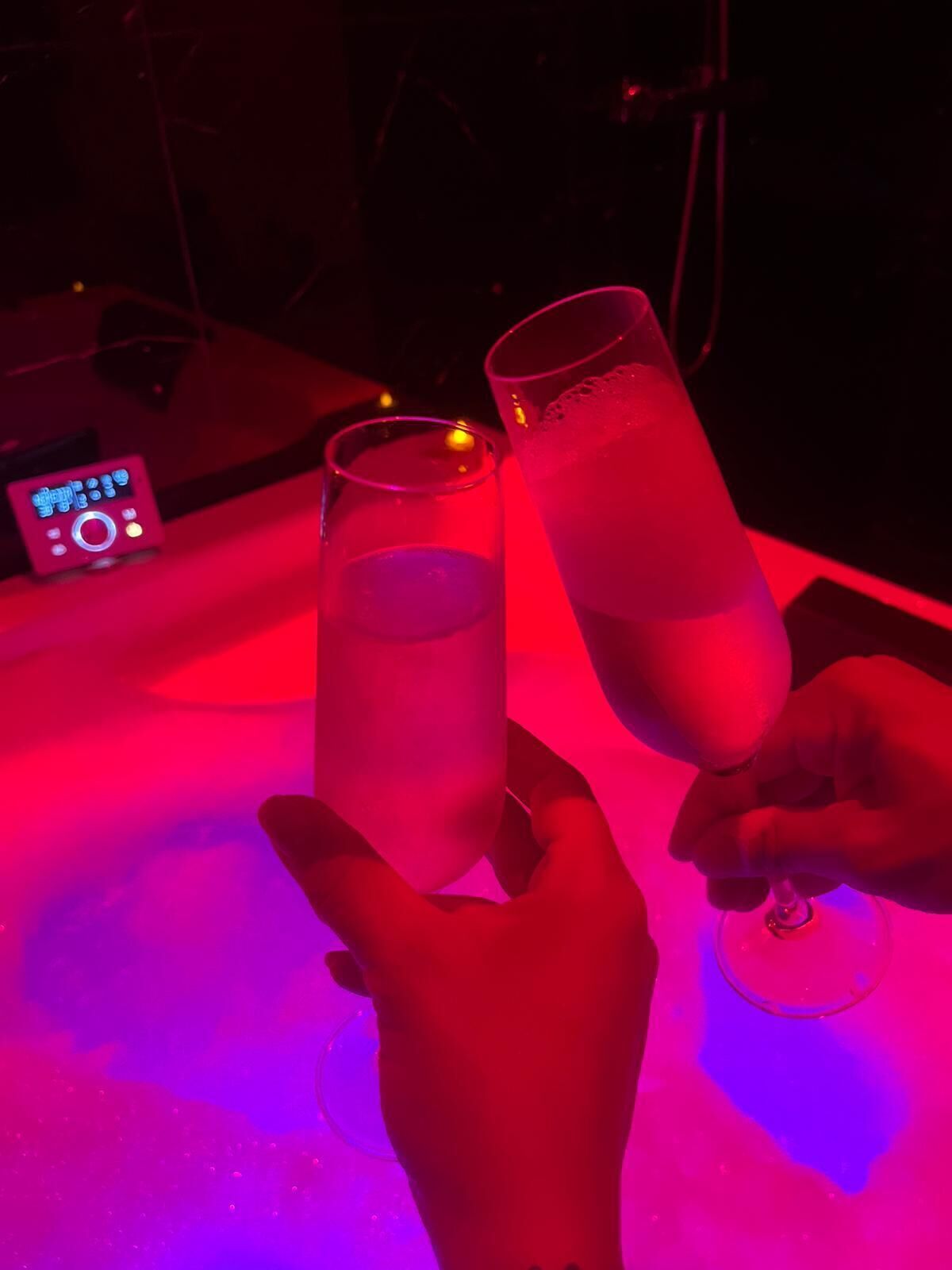 Two people holding tall glasses of a clear liquid, likely a beverage, in a dimly lit setting with a red hue.