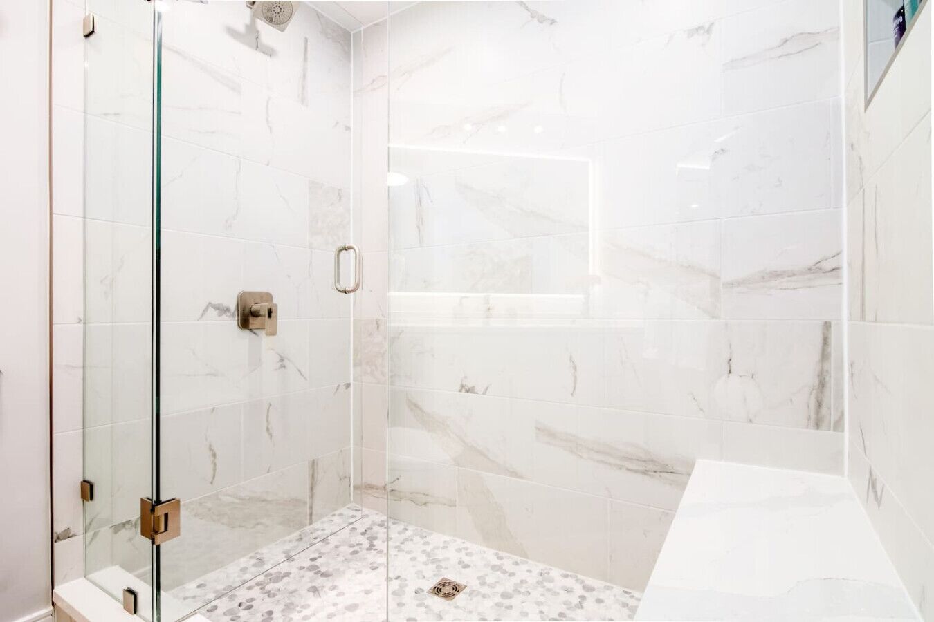 Une salle de bain moderne avec une douche à l’italienne en verre, des carreaux de marbre et un plan de toilette blanc.