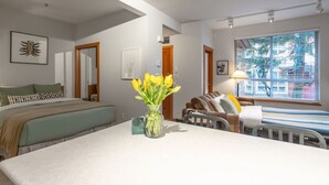 Habitación de hotel con cama, escritorio con un florero y ventana con persianas, con vistas a árboles.