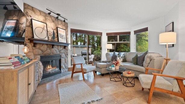 A cozy living room with a stone fireplace, comfortable seating, and large windows offering a view of greenery.