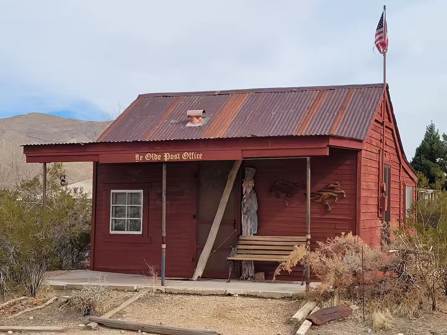 Un bâtiment rustique de couleur rouge, portant l’inscription « Je Olde Post Office », avec un toit en métal, un banc en bois à l’extérieur et un drapeau américain sur un mât.