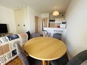 Espaço de estar compacto com uma mesa de jantar redonda de madeira, duas cadeiras cinzentas, um sofá estampado, uma televisão e uma cozinha compacta ao fundo.