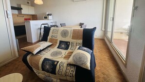 Un salon chaleureux avec un canapé à motifs, un coin repas avec une table et des chaises, une cuisine équipée de placards et d’appareils électroménagers, et une grande fenêtre offrant une vue dégagée.