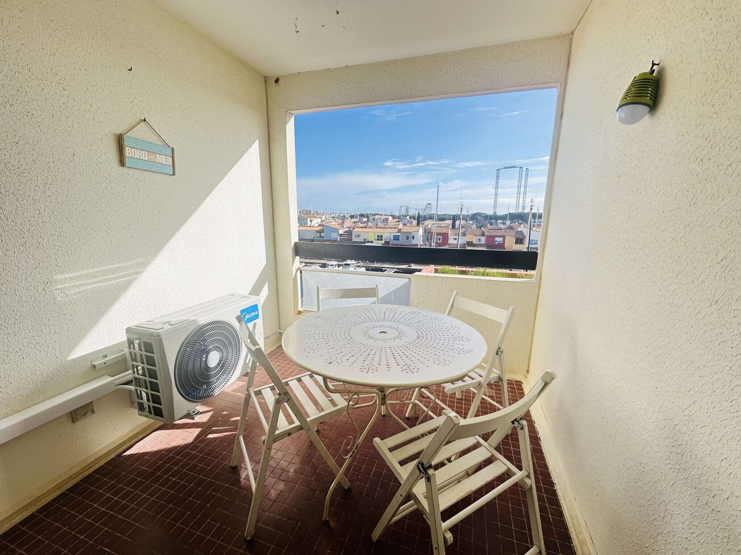 A small balcony with a white table and chairs, a fan, and a view of a cityscape.