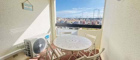 A small balcony with a white table and chairs, a fan, and a view of a cityscape.