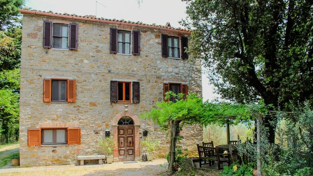 A stone building with a central door, multiple windows with shutters, and a garden area with outdoor furniture.