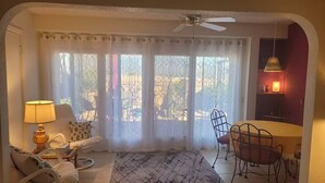 Un salon comprenant un canapé, une table à manger avec des chaises, un tapis à motifs, un ventilateur de plafond et une vue sur l’extérieur à travers une fenêtre avec des rideaux.