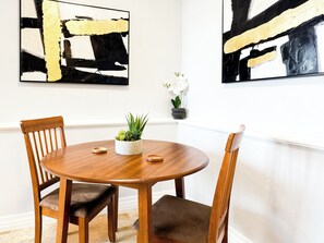 A dining area with a round wooden table, two wooden chairs, a potted plant, and two abstract paintings on the wall.