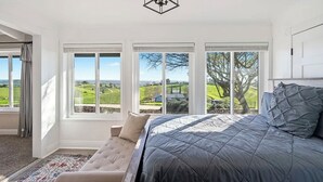 A bedroom with a large bed, a bench, and three windows offering a view of a green landscape.