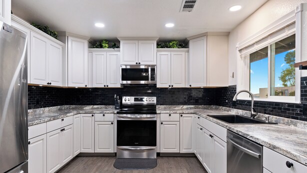 A modern kitchen with white cabinets, a black backsplash, stainless steel appliances, and a window with blinds.