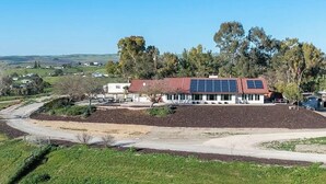 A building with a red roof and solar panels, surrounded by greenery and a dirt path.
