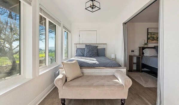 Ein Schlafzimmer mit Bett, Sessel, Nachttisch und Fenster mit Blick auf Grünflächen.