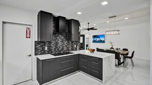 A modern kitchen with dark cabinetry, a tiled backsplash, and a dining area with a TV displaying 'MIAMI' on the wall.
