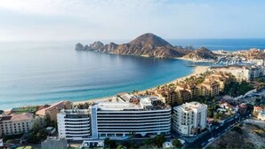 A coastal cityscape with a prominent hotel featuring multiple floors and balconies, a beach, and a mountainous backdrop. - New luxury resort on Medano Beach, (Cabo San Lucas)