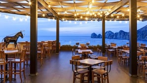 An outdoor dining area with wooden tables and chairs, string lights, and a view of the sea and mountains. - New luxury resort on Medano Beach, (Cabo San Lucas)
