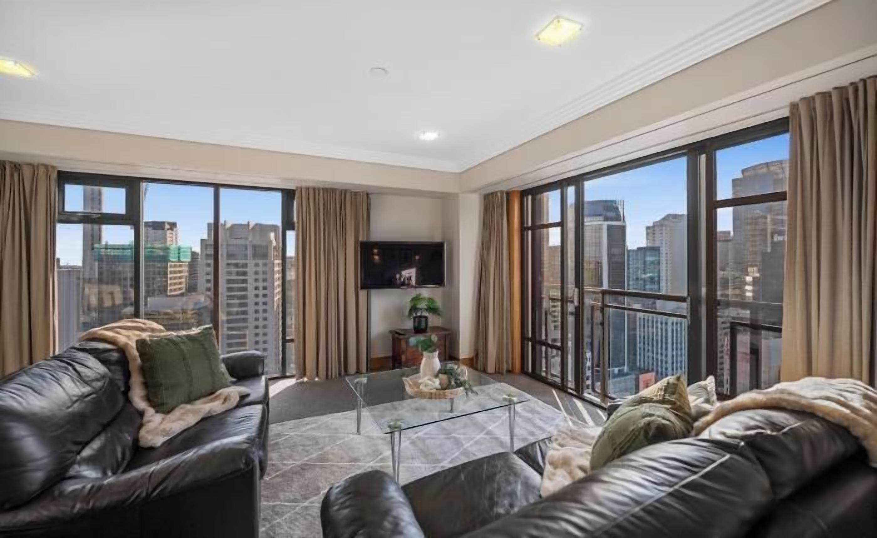 A spacious living room with a leather sofa, a glass coffee table, and a flat-screen TV mounted on the wall. Large windows offer a view of the cityscape.