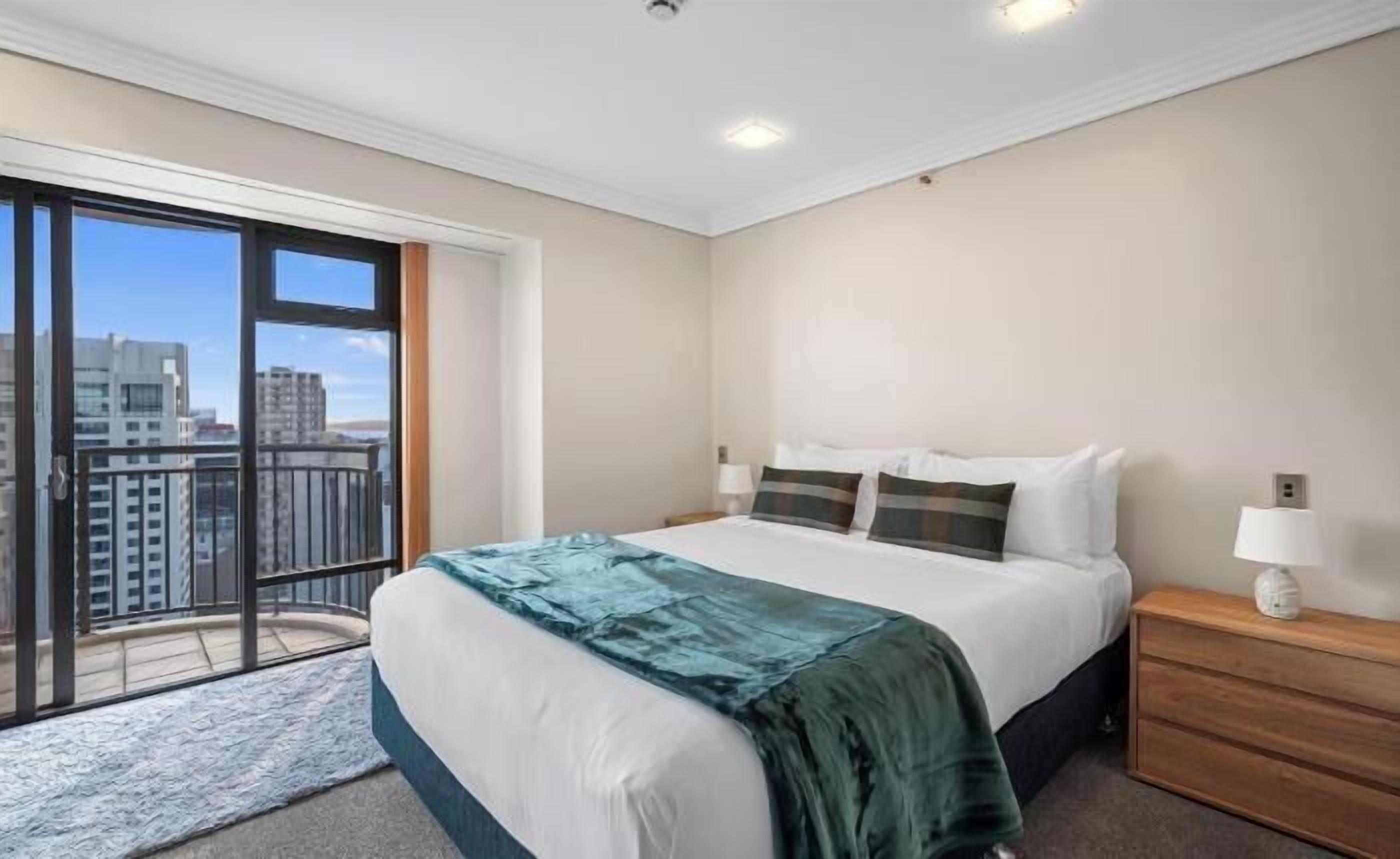 A bedroom with a large bed, a nightstand, a lamp, and a view of the city through a balcony.
