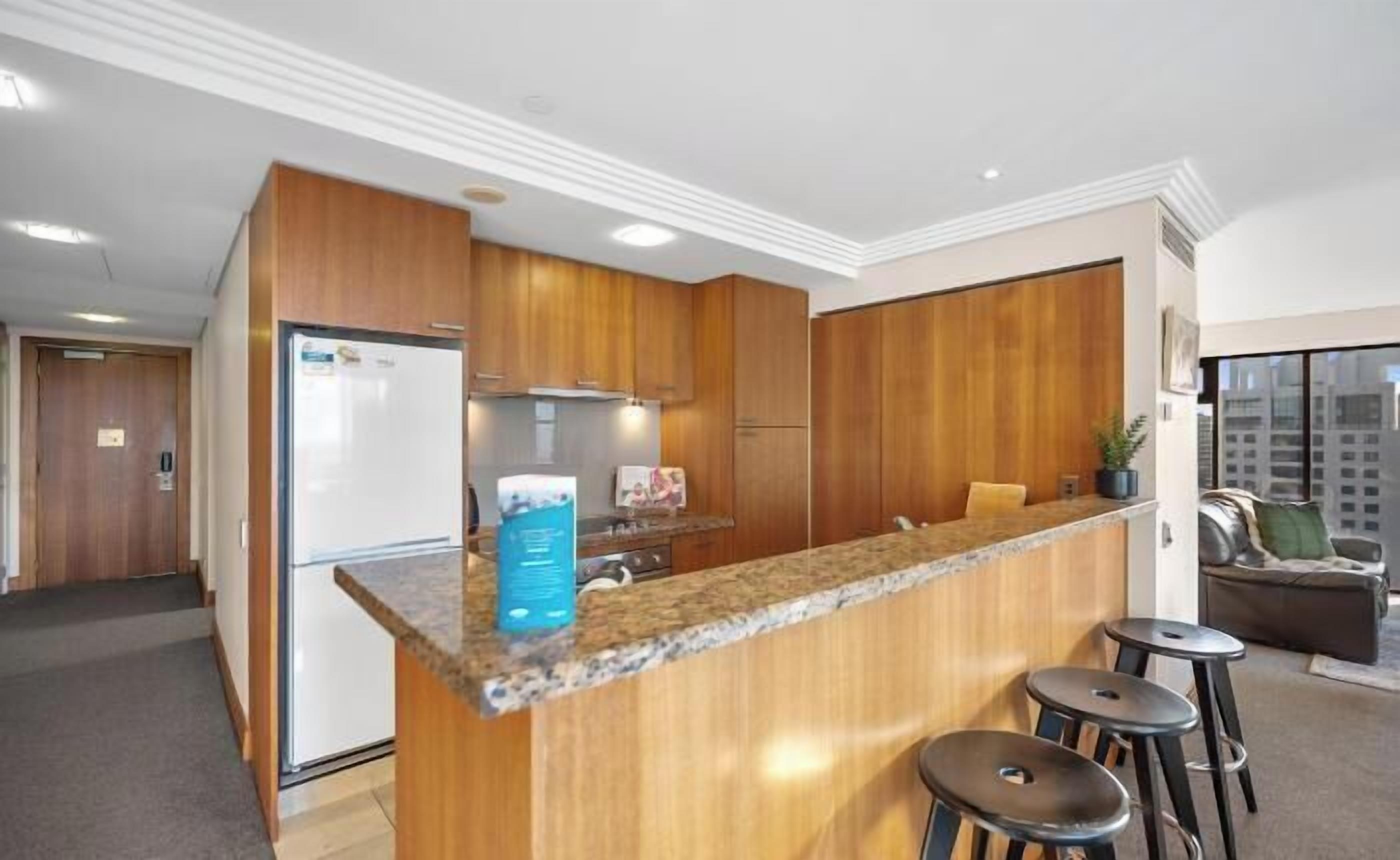A modern kitchen with wooden cabinets, a granite countertop, and a built-in refrigerator. There are two bar stools and a large window offering a city view.