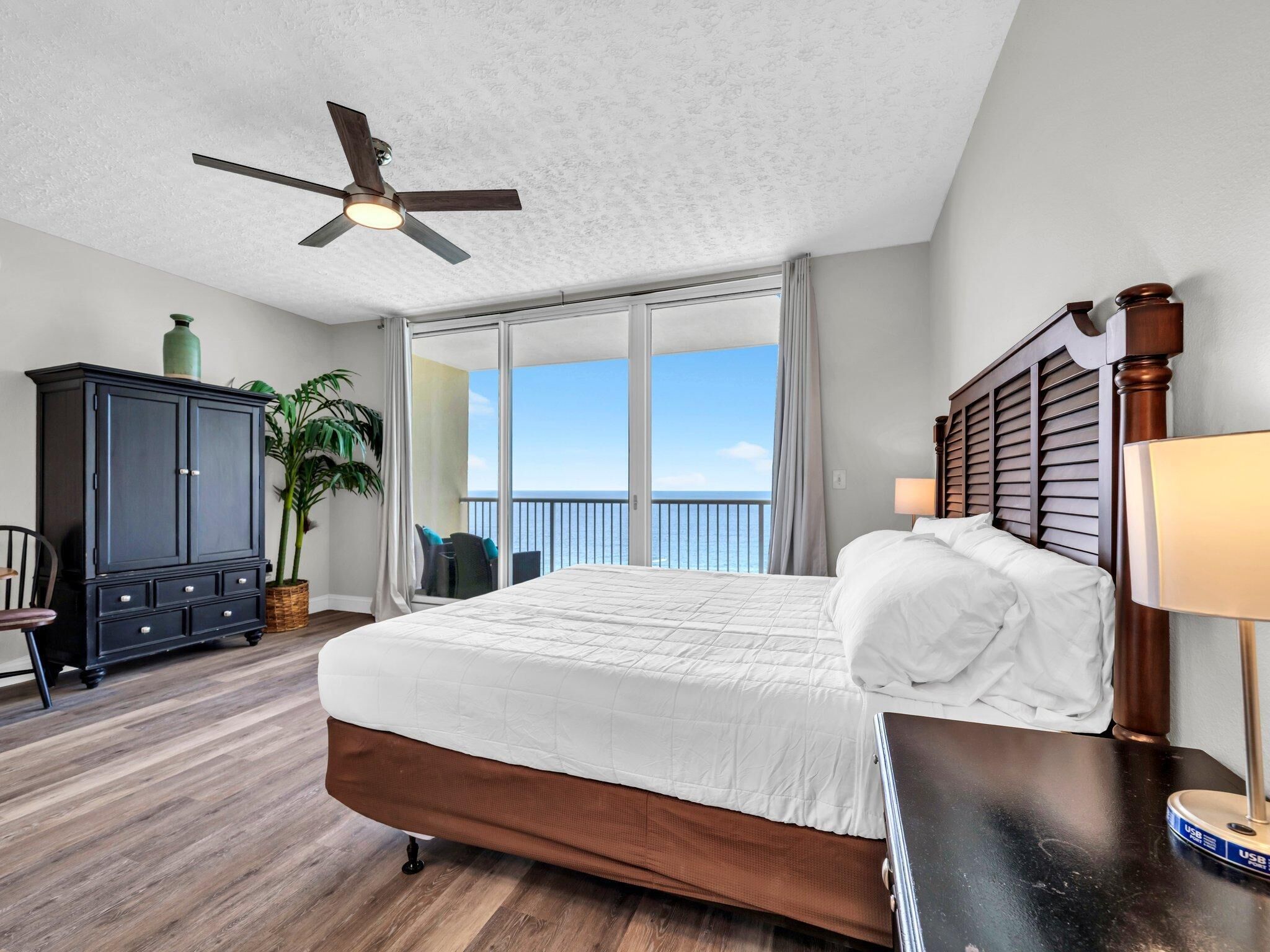 A hotel room with a bed, a ceiling fan, a dark wooden cabinet, a plant, and a view of the ocean through large windows.
