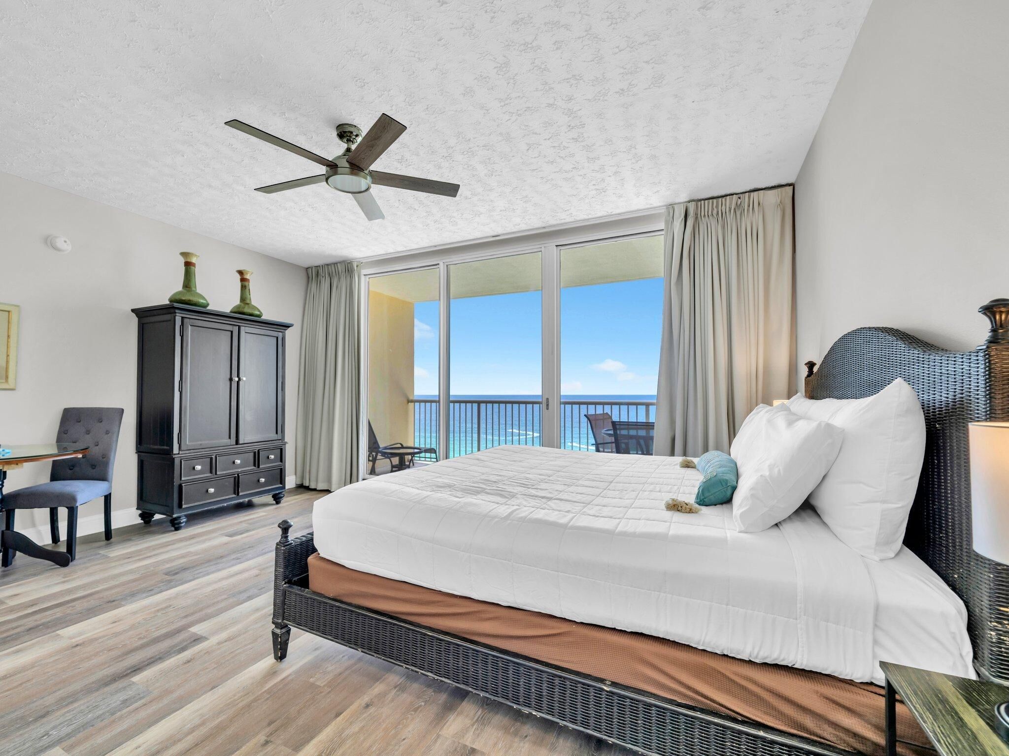A hotel room with a large bed, a ceiling fan, a view of the ocean, a balcony with a table and chairs, and a dark wooden wardrobe.