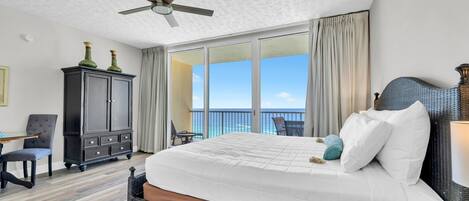 A hotel room with a large bed, a ceiling fan, a view of the ocean, a balcony with a table and chairs, and a dark wooden wardrobe.