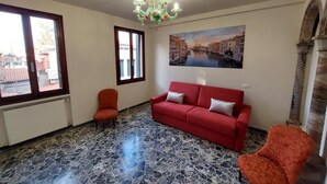 Quarto com sofá vermelho, duas poltronas, um candelabro, pintura de um canal e chão de pedra.