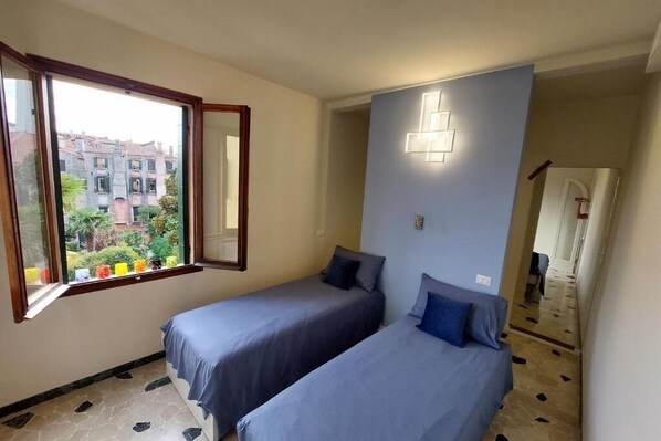 A bedroom with two single beds, a window with a view of buildings, and a wall-mounted light fixture. - Donini Residence in Venice, Suite 1 (venezia)