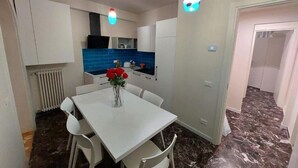 Uma cozinha compacta com armários brancos, uma parede de azulejos azuis e uma área de refeições com mesa e cadeiras brancas.