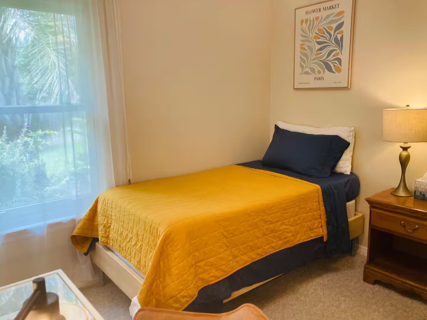 Un dormitorio con cama, una mesita de noche con lámpara, un cuadro en la pared y una ventana con vistas a zonas verdes.