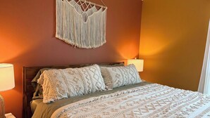 A bedroom with a bed, two bedside lamps, a macrame wall hanging, and a bedside table. - Rest In Peace on Perkins (San Marcos)