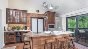 A kitchen with wooden cabinets, a central island with stools, a built-in oven, and a large window offering a view of greenery. - Little Sandy Cove Paradise (Siesta Key)