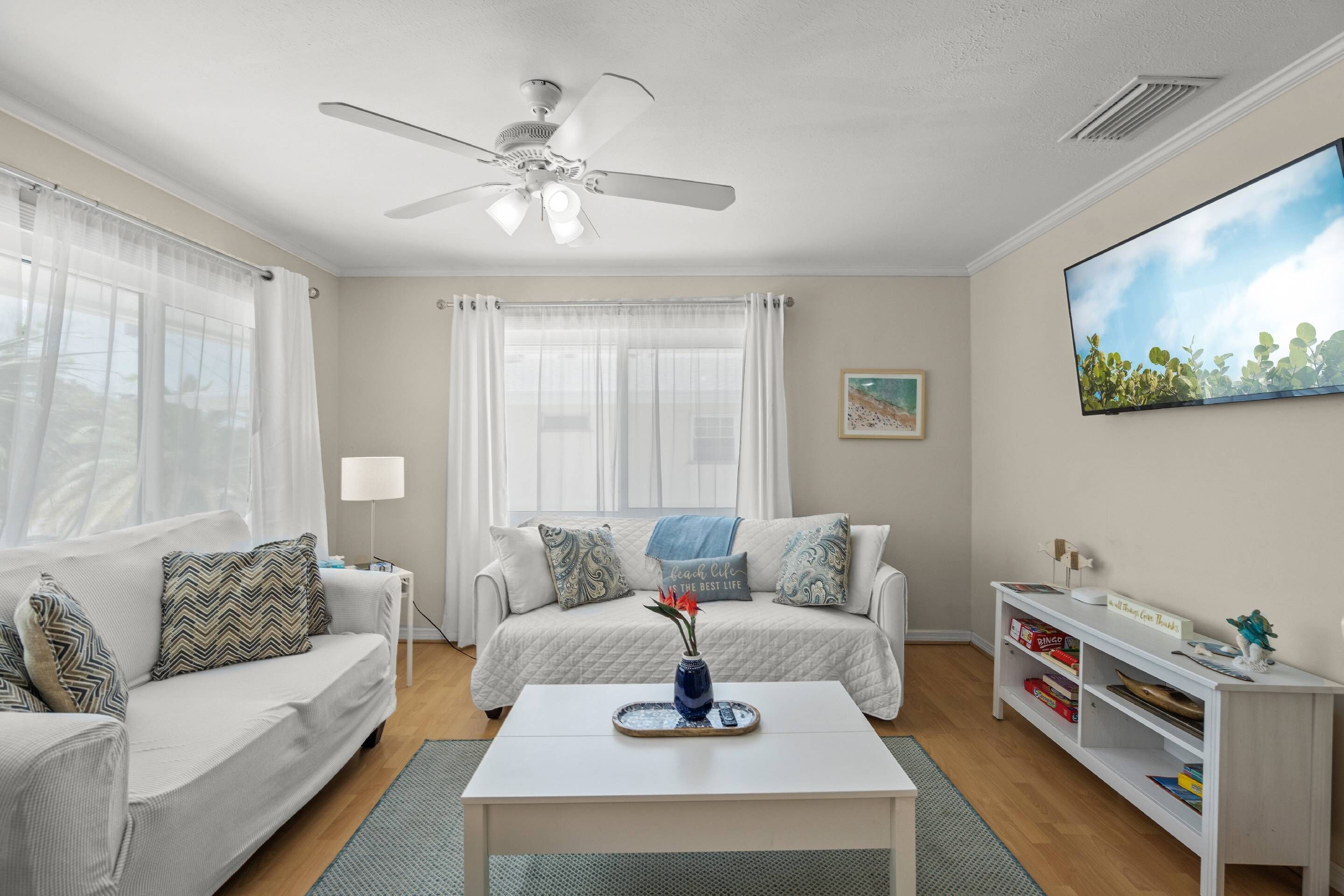 A room with a white ceiling fan, a white sofa with patterned cushions, a white coffee table with a vase, a white TV stand with a flat-screen TV, and a bookshelf.