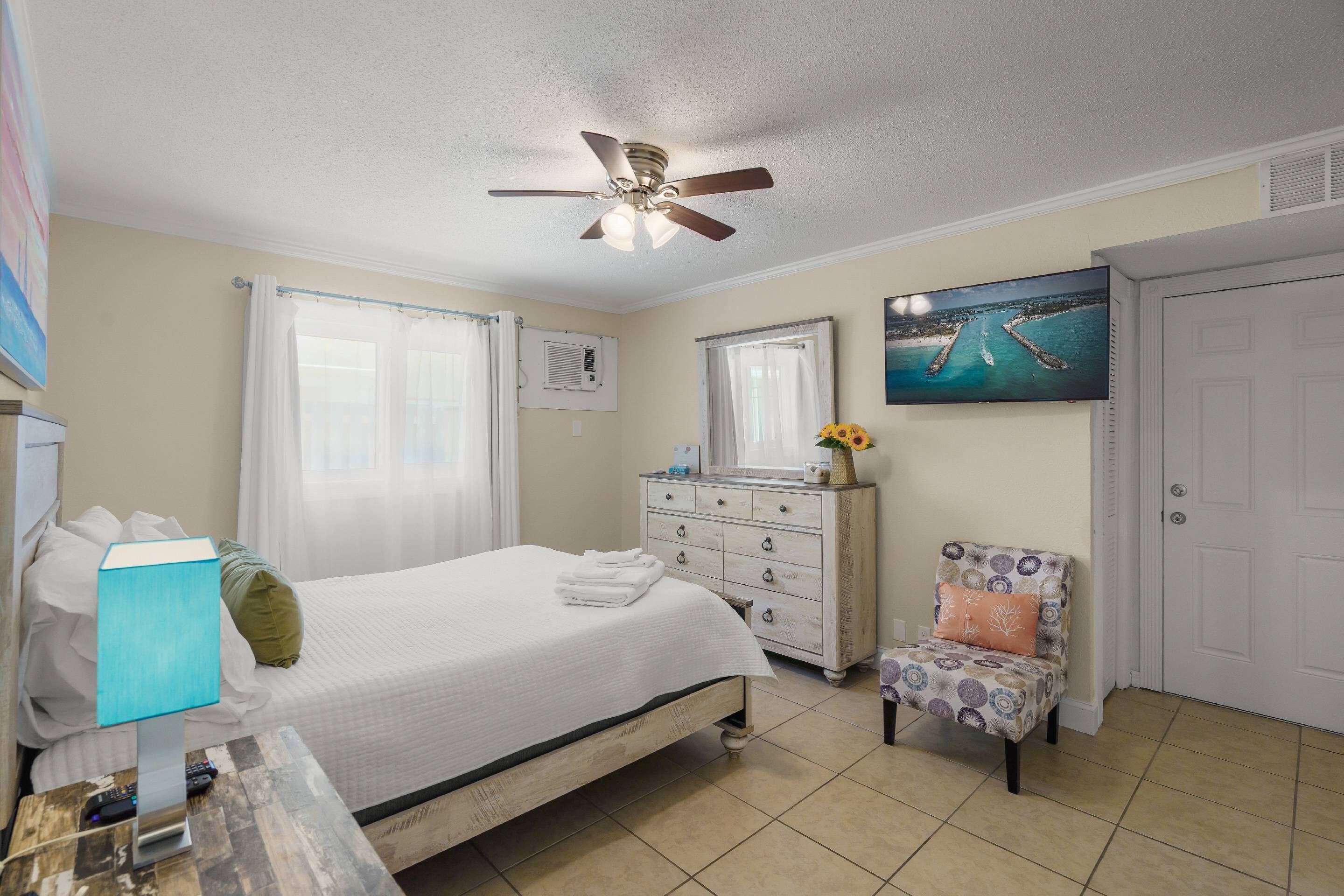 A bedroom with a bed, a dresser, a chair, a ceiling fan, a painting, and a door.