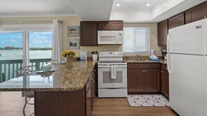 A kitchen with granite countertops, a white stove, a microwave, and a refrigerator. There is a dining area with a table and chairs, and a view of the water outside. - Coastal Breeze Siesta - Pelican (Siesta Key)